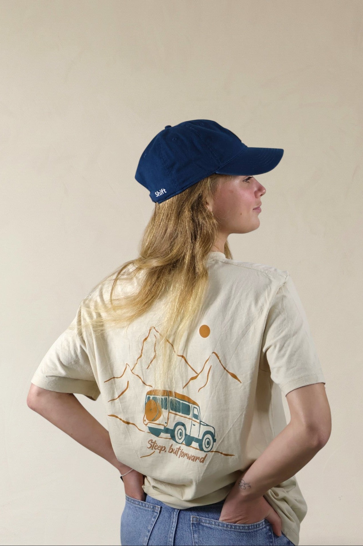 Person wearing a blue cap and a beige t-shirt with a graphic design on a plain background