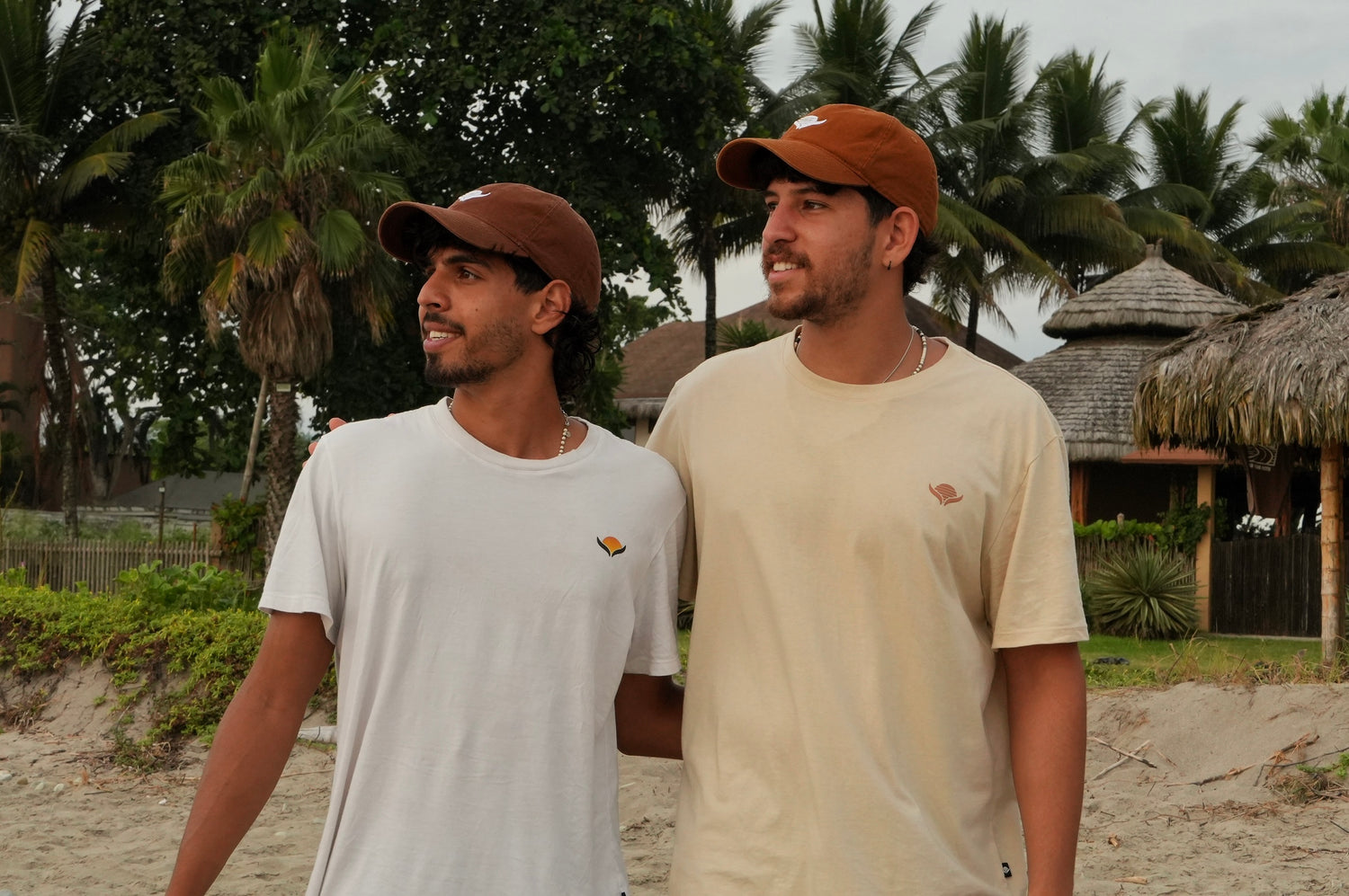 Two men wearing brown hats and t-shirts on a beach with palm trees in the background.