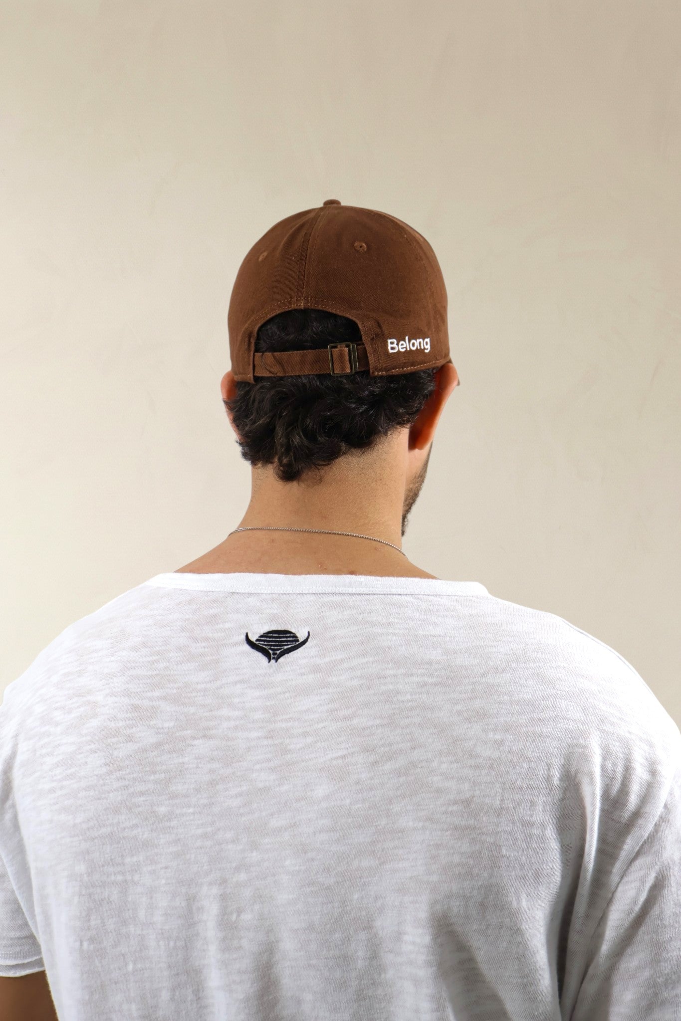 Person wearing a brown cap with 'Belong' logo and white t-shirt with black logo on beige background