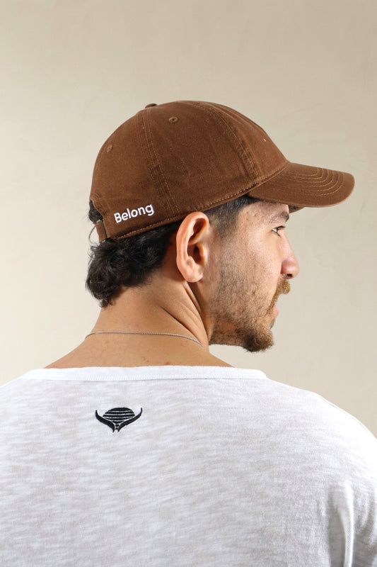 Man wearing a brown cap with 'Belong' text and a white t-shirt with a black logo on a beige background