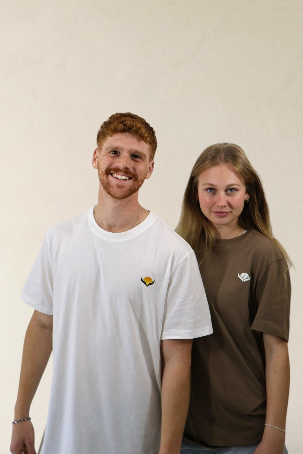 Two people wearing white and brown t-shirts with small logos against a plain background