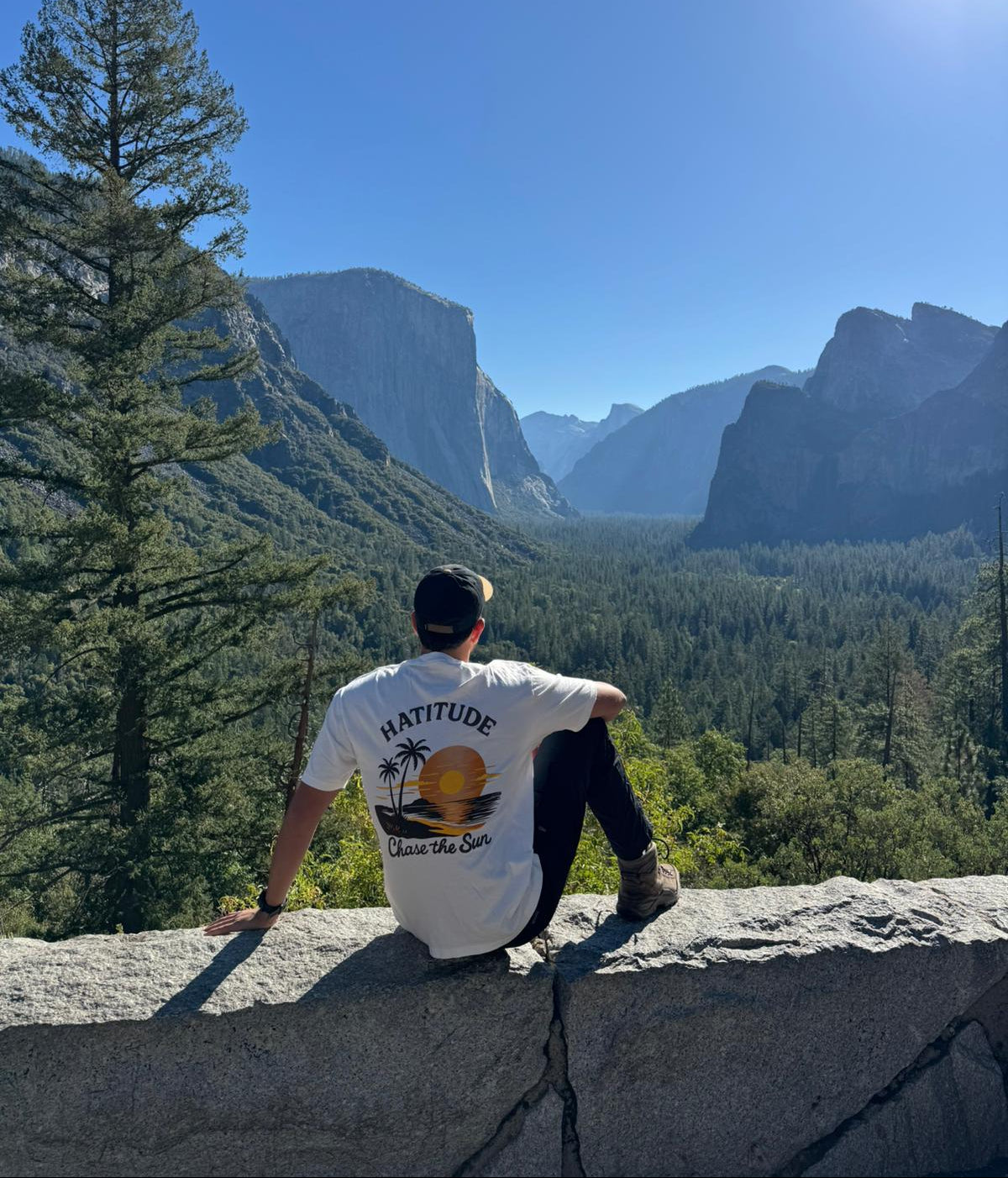 Person sitting on a ledge overlooking a mountainous landscape with a clear blue sky.
